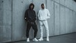 © Exclusive  - In an urban environment, two young men stand confidently against a textured concrete wall, wearing contrasting hooded sweatshirts and casual athletic wear