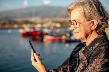 Smiling senior woman in casual jacket using mobile phone at sea port in sunset light. Elderly relaxed woman holding her smartphone enjoying tech and social