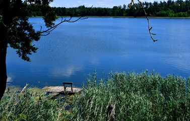 A view of a vast river or lake surrounded from all sides with reeds, forests, moors, fields, and pasturelands located next to a small marina or wooden platform and some metal tower seen in Poland