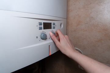 The photo shows a woman's hand with long nails turning the control knob on the gas boiler panel to set the temperature in a private home.