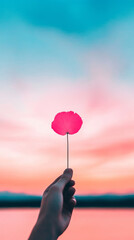 Sticker - A hand holds a single pink flower against a vibrant sunset sky.