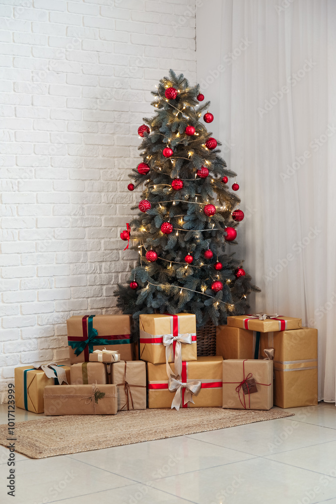 Decorated Christmas tree with balls, glowing lights and gift boxes near white brick wall
