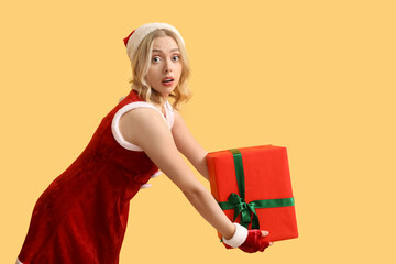 Canvas Print - Emotional young woman dressed as Santa Claus with Christmas gift on yellow background