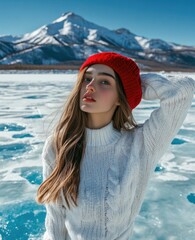 Poster - A beautiful girl wearing a white sweater and red hat, posing on a frozen lake with a snow-capped mountain background.