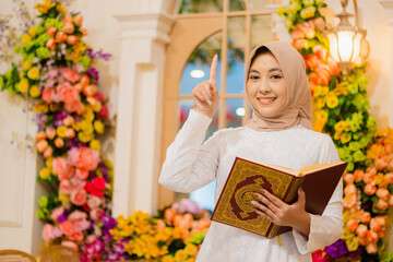 girl hijab wearing a white dress muslim cheerful to the camera both hands holding a book, the quran is open while pointing upwards in a luxurious interior. for fashion, promotion, lifestyle, religious