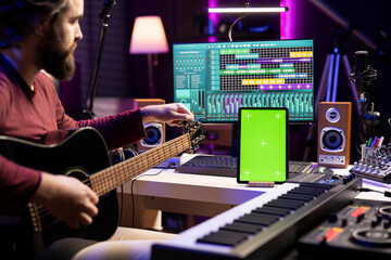 Leinwandbilder - Musical engineer learning to tune his acoustic guitar before playing, watching online video tutorial by using greenscreen on tablet. Young songwriter producer tuning accords on instrument.