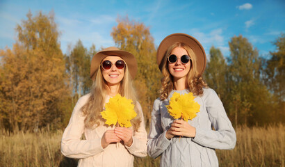 Wall Mural - Happy young two women friends with autumn yellow leaves, stylish girlfriends smiles together in park