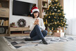 © sofiko14 - Young Caucasian woman wearing Santa hat sitting in living room near Christmas tree using smartphone. Cozy holiday atmosphere with decorated tree, wrapped gifts, and festive decor.