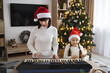 © sofiko14 - Caucasian mother and daughter wearing Santa hats play electronic piano together during Christmas. Warm family moment with girl and parent celebrating holiday, creating festive atmosphere.