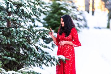 Woman in a red lace dress touches snowy pine branches in winter.