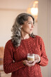 © Cavan Images - Woman looking out a window at her home holding coffee