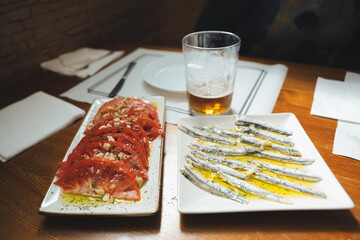 Sliced tomatoes and anchovies elegantly arranged as tapas on a rustic wooden table with a glass of beer. Enjoy these delectable tapas in a cozy restaurant setting