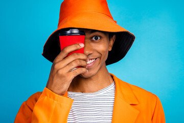 Poster - Portrait of happy guy smiling hide face with to go coffee mug wear stylish hat isolated blue color background