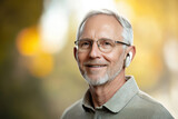 Senior Man Wearing Wireless Earbuds and Glasses Outdoors, Smiling older man with gray hair, glasses, and wireless earbuds enjoying an outdoor setting with a blurred background