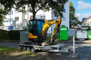 A yellow excavator mounted on a trailer is parked beside residential buildings, with green trash bins and trees in the background, showcasing a clear and sunny day.