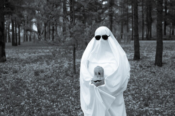 Woman in white sheet holding jack o'lantern against autumn forest background. Halloween costume. Black and white photo. 