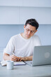 © wifesun - Focused man in glasses and white t-shirt working at home with laptop, writing in notebook, doing remote work or study sitting in bright kitchen with mug of coffee, minimalist design