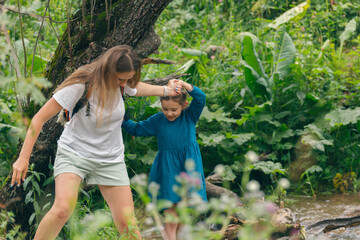 A mother assists her daughter in navigating a lush, green forest stream, fostering a connection with nature.