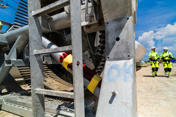 Two engineers in safety gear inspecting a wind turbine blade section on a construction site. They examine the metal framework with precision. Renewable energy project under a clear blue sky.