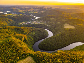 Golden hues illuminate the Vltava River as the sun sets behind rolling hills in Czechia, creating a breathtaking view of the water winding through lush forests and serene landscapes.