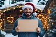 ©  S u n n y B u n n y - Cheerful man in santa hat holding blank cardboard sign in festive winter setting.