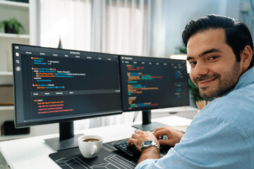 Smiling smart IT developer working software development looking camera with coding on two pc creating virus firmware application program version on website with coffee cup at modern office. Surmise.
