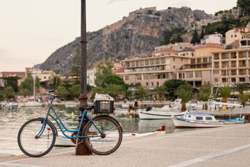 A charming blue bicycle rests by a lamppost along the tranquil Nafplio waterfront. As the sun sets, boats float peacefully in the harbor, creating a serene atmosphere.