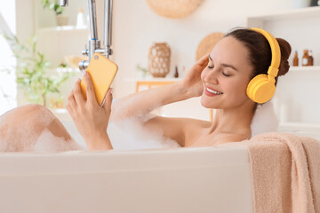 Wall Mural - Beautiful young happy woman in headphones holding mobile phone and taking bath with soap foam at home