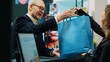 © DC Studio - Checkout staff selling clothing items to senior customer in shopping mall, preparing clothes in a paper bag for client. Elderly man approaching cash register to buy merchandise. Camera A.