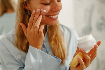 A cheerful woman is happily enjoying her skincare routine by gently applying her favorite moisturizer to her face