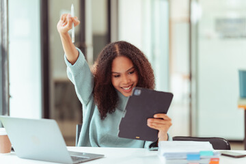 Happy young African woman with curly hair and raised arms in victory pose in front of laptop at business goal, project completed.