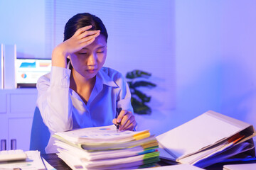 A young woman works overtime at night at her desk in office, using a laptop reviewing documents. is stressed, tired,experiencing headaches,body aches from sitting too long,leading to office syndrome