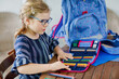 © Irina Schmidt - Happy smiling girl preparing for school her backpack. First day of school. Back to school concept. Little child collecting different supplies like pens and books in a bag or satchel.