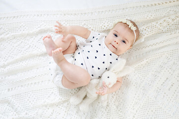 Wall Mural - a baby girl of six months on the bed at home playing with a soft toy and her legs, a smiling little child with a toy in her hands on the bed in the bedroom