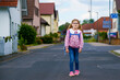 © Irina Schmidt - Happy little girl with glasses, wearing a pink sweater and a blue backpack walking to elementary school. She is smiling confidently, ready for school on a pleasant day.