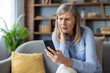 © Liubomir - Mature woman sitting on sofa looking worried at smartphone. Facing technology issue or confusing news, expressing concern and focus. Casual setting at home, adult in blue shirt.