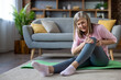 © Liubomir - Senior woman with knee pain sits on floor while exercising at home, expressing discomfort and concern. Relaxing in living room, she rests on green yoga mat, wearing casual attire.