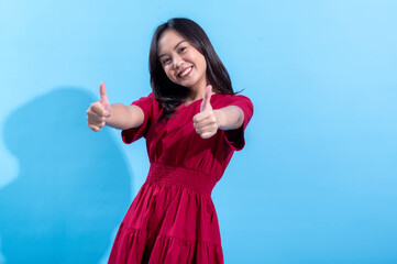 A cheerful young Asian woman in a bright red dress stands against a light blue background. She is smiling widely and giving two thumbs up, expressing enthusiasm and positivity