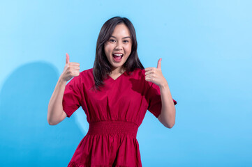 A cheerful young Asian woman in a bright red dress stands against a light blue background. She is smiling widely and giving two thumbs up, expressing enthusiasm and positivity