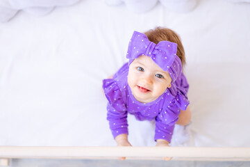 cute baby girl in a crib is holding on to the side in lilac clothes and with a bow on her head, funn