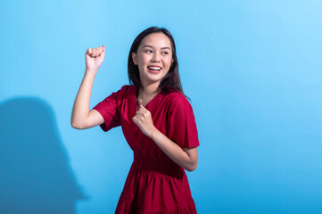 An excited Asian woman in a red dress raises both fists in the air while cheering enthusiastically. She stands against a light blue background, displaying an expression of joy and triumph