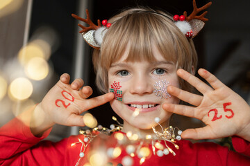 cheerful child holds his palms near face on which numbers of new year 2025 are written. boy is waiting for miracle on Christmas Eve. winter holidays at home. festive pictures