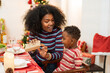 © winnievinzence - Excited mother unwrapped gift box for her son at dining table. African family enjoy celebration Christmas festive dinner on holidays together at home with xmas decoration theme