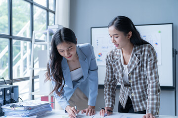 Two asian businesswomen are discussing and working together over paperwork at a desk in a modern office. One of them is writing on a document