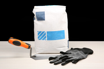 Wall Mural - Bag of cement powder, putty knife and rubber gloves on light wooden table against black background. Building material