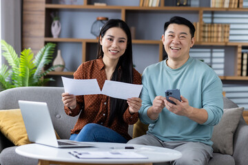 Cheerful asian couple trusts each other reviewing financial documents with phone and laptop. Together they plan budget with teamwork, happiness, and technology in home setting.