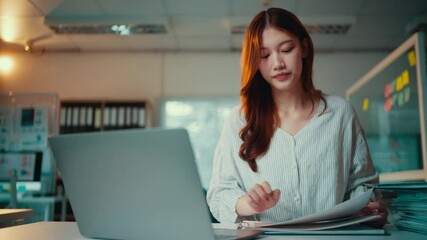 Wall Mural - Asian accountant reviews financial documents while working on a laptop in a professional office environment, focusing on data analysis and financial planning tasks