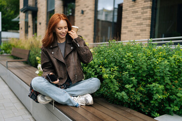 Wall Mural - Joyful young redhead female in leather jacket sitting on urban bench, enjoying cup of coffee and using mobile phone, taking break from day to relax and catch up with friends on summer day outdoors.