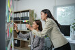 © amnaj - Two businesswomen are having a discussion about a new project, writing ideas on sticky notes and sticking them on a glass wall in the office