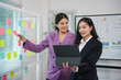 © amnaj - Two businesswomen are having a discussion in an office, using a tablet and sticky notes on a glass wall to brainstorm ideas